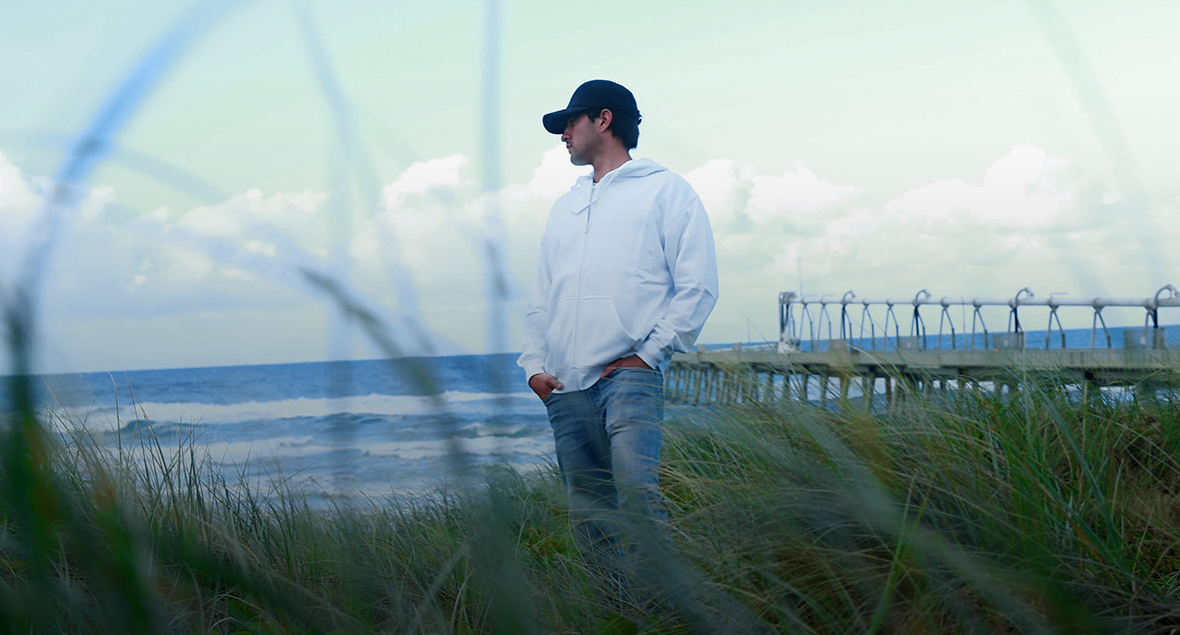 Man in white hoodie and INIVI cap by ocean.